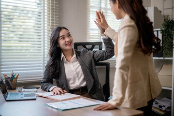 Professional women high-fiving, sharing workplace achievement near computer workstation