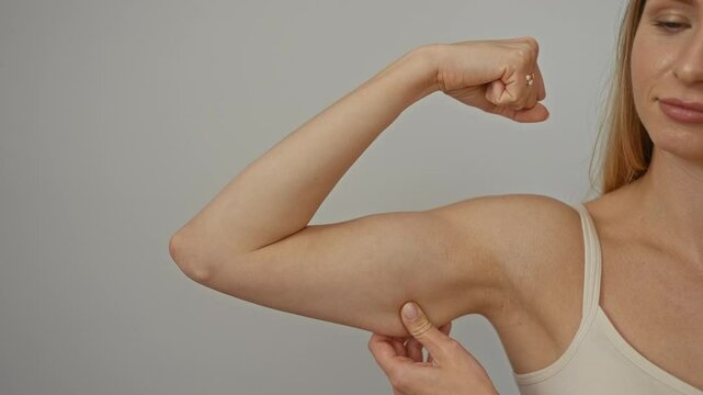 Woman flexing arm muscle while pinching skin on isolated white background, showcasing strength and body awareness