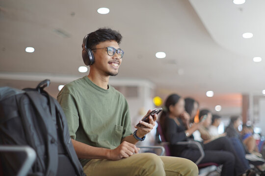 Smiling young Asian man using smartphone and headphones, chatting online, listening to music, while waiting in the waiting area and waiting to check in. - Powered by Adobe