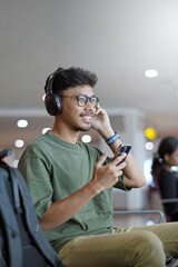 Smiling young Asian man using smartphone and headphones, chatting online, listening to music, while waiting in the waiting area and waiting to check in.