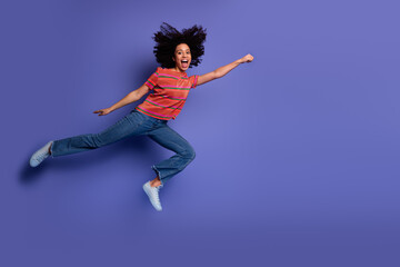 Cheerful young woman leaping joyfully in a colorful outfit against a purple background expressing...