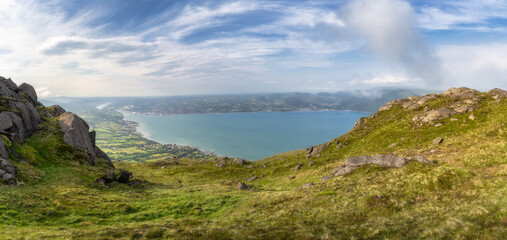 A stunning panoramic view that showcases the lush, vibrant green hills beautifully meeting the calm, tranquil blue waters beneath a picturesque, cloud filled sky, Carlingford, Ireland