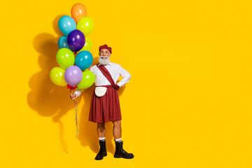 Cheerful elder man dressed in traditional scottish kilt holding colorful balloons, standing on vibrant yellow background