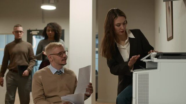 Young female office worker scanning papers on corporate scanner while printing out documents cooperation with male colleague using wheelchair captured in hallway
