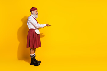 Mature man in traditional Scottish kilt gesturing against bright yellow background highlighting cultural attire and charm