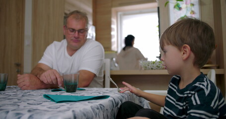 Obraz premium Father looking at son at the table during a family moment in a kitchen setting, while the mother works in the background, highlighting connection and routine