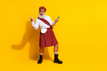 Mature man in traditional Scottish attire cheerfully using a smartphone on a yellow background exuding positive energy