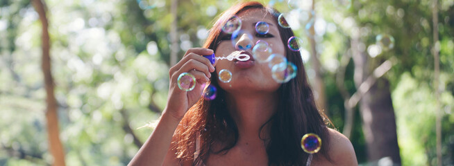 Young woman joyfully blows soap bubbles outdoors with sunlight and greenery background, carefree and playful summer moment in nature-banner