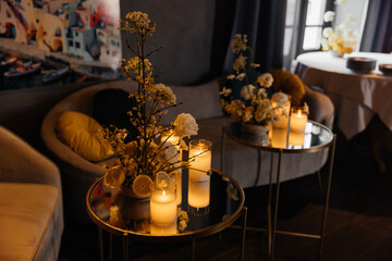 A composition of candles, flowers, and lemons on small tables with armchairs in the background, set in a modern interior. Home decor.