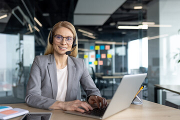 A friendly woman in a headset smiles while working on her laptop in a modern office setting. She...