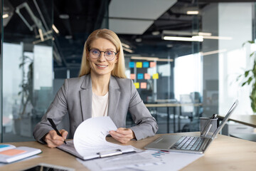 A smiling businesswoman, wearing glasses, sits at her desk in a modern office. She is reviewing documents, ready to sign. The laptop is open.