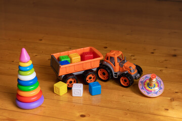 Fototapeta premium A variety of colorful plastic toys are scattered on a wooden floor, including a stacking ring pyramid, a toy dump truck with a trailer, multicolored building blocks, and a spinning top.