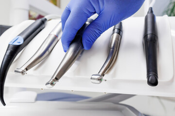 A gloved hand selects a dental handpiece from a set of tools on a white holder. The clinical setting features neatly arranged instruments and a clean background.