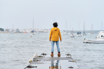 Man wearing yellow raincoat standing on pier looking at sailboats in harbor on a cloudy day