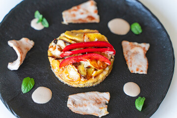 Slices of cooked turkey with potato tortilla and red bell peppers on a black plate with sauce. Close-up, top view.