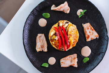 Slices of cooked turkey with potato tortilla and red bell peppers on a black plate with sauce. Close-up, top view.