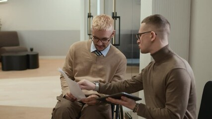 Medium shot of male office worker in wheelchair talking to colleague discussing contract terms and conditions while meeting during working day in hallway