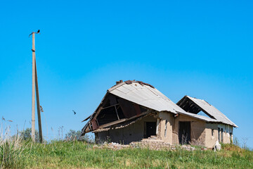 Obraz premium A close-up of two dilapidated one-story brick houses and a pole without wires, standing in the middle of a meadow against a blue sky. A common Roller or raksha (Coracias garrulus) bird hunts nearby.