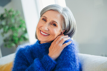 Grey haired woman in blue sweater smiling warmly indoors, enjoying natural daylight and a peaceful...