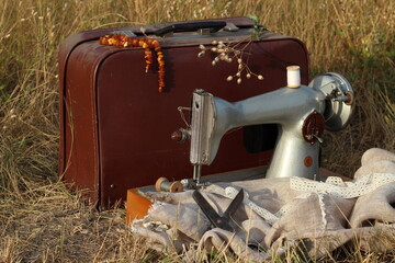 sewing machine with a brown suitcase on a background of dry grass with an old pair of scissors and...