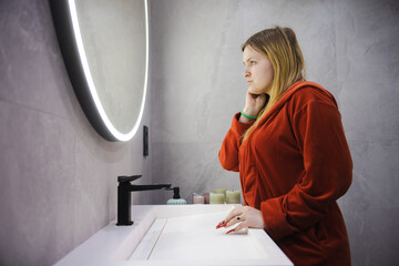 Young woman in a red bathrobe leans against the sink, resting her face on her hand with a weary expression while staring into a glowing round mirror.