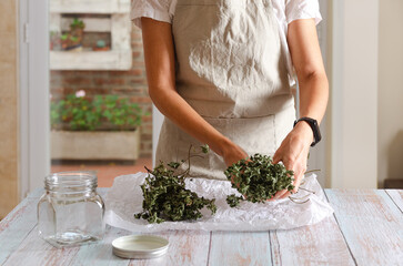 A woman with a beige apron separating dried mint leaves from the branches on a white paper for storing in a jar. 