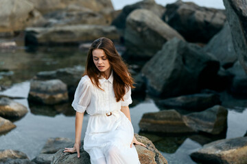 Serene woman in white dress sitting on ocean rock with rocky background
