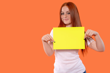 Naklejka premium Young woman with red hair holds bright yellow paper blank against vibrant orange background while smiling and pointing down