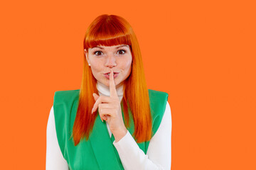 Woman with vibrant red hair gestures for silence against a bright orange background, creating an...