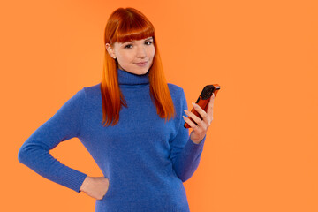 Young woman with bright red hair smiles while holding a smartphone against an orange background in...