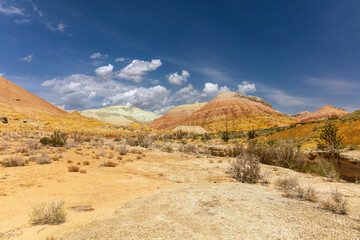 Vibrant multicolored mountain landscapes with red, yellow, and white layers on the hillsides, contrasted by desert vegetation in the foreground. A bright blue sky with fluffy white clouds accentuates.