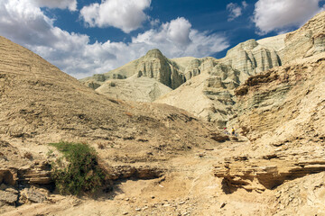 Breathtaking arid landscape featuring rugged beige rock formations and eroded cliffs under a vivid blue sky with scattered clouds. The scene includes two cyclists navigating through the canyon, adding
