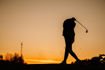 Silhouette of a female golfer completing a powerful follow-through at sunset, with debris in the air and golf bag resting nearby under orange sky..