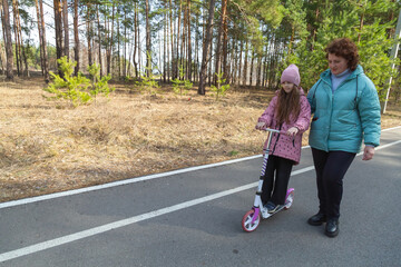 Little girl riding scooter while walking with her grandmother in the forest on a sunny day