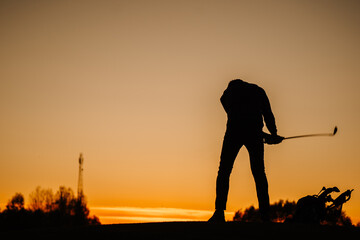Silhouette of a golfer bending with club in hand at sunset, next to a golf bag, set against a...