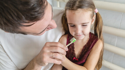 A loving father is holding his daughter in the arms and helping her brushing their teeth in a...
