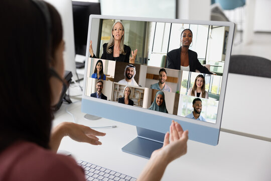 Conference call. Over shoulder view young businesswoman in earphones with mic talk to diverse partners group on remote business event before pc screen leading workshop training in corporate video chat