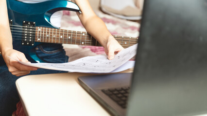 Fototapeta premium Young caucasian woman using laptop while sitting on bed and playing electric guitar at home