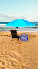 Mobile photo of the tranquil beach scene with a blue beach umbrella providing shade over a lounge chair and a striped deck chair. The calm ocean waves gently lap against the sandy shore.