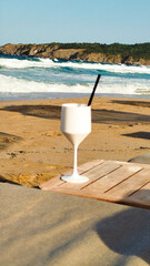A picturesque beach scene featuring a white glass with a drink and a straw on a wooden table. The clear blue sky and gentle waves offer a perfect backdrop for a day of relaxation by the sea.
