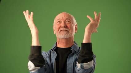 Naklejka premium A mature man with a gray beard displays intense emotions, raising his fists in excitement or celebration. He stands against a solid green backdrop.