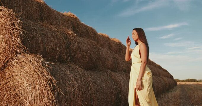 Fashion model posing in a yellow dress by a haystack in a field at sunset