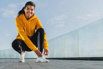 Young fitness woman tie shoelaces on road runner sitting on floor on city streets in earphones...