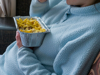 Relaxed woman lounging in pajamas savoring kebab and french fries from aluminum take away container while resting comfortably on home sofa