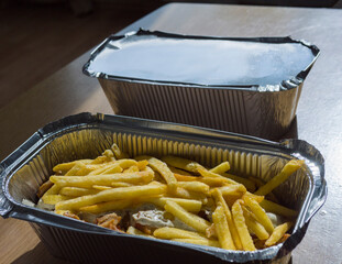 French fries covering a kebab meat with white garlic sauce in an open aluminum take away box on a wooden table, with another closed aluminum box next to it