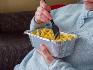 Relaxed woman lounging in pajamas savoring kebab and french fries from aluminum take away container while resting comfortably on home sofa
