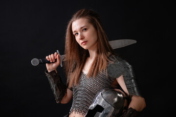 Portrait of a medieval female warrior in armor holding a sword and helmet in her hands, posing standing against a dark background.