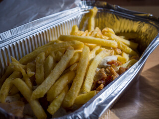 French fries piled on a kebab drizzled with sauce, all nestled in an aluminum takeaway box resting on a wooden table, creating a tempting fast food meal