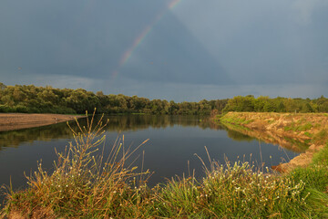 A summer landscape depicting a wide river with a rainbow over the river