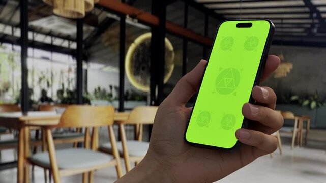 GREEN SCREEN CHROMA KEY Person holding a smartphone in a cozy cafe with modern interior design, wooden furniture, and natural daylight ambiance